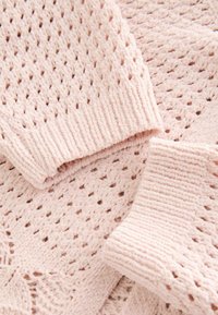 Tissu tricoté rose doux avec des motifs ajourés complexes et des poignets côtelés, probablement provenant d’un pull ou d’un cardigan.