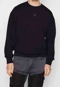 Sudadera negra hecha de un tejido suave, con cuello redondo y un pequeño logo en relieve en la parte delantera. Diseño holgado.