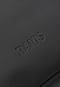 Svart vattentätt material med en slät yta, prytt med en präglad "RAINS"-logotyp. Enkel design, minimalistisch utan ytterligare detaljer eller mönster.