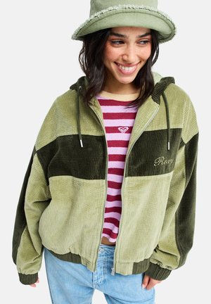 Młoda kobieta uśmiechająca się, ubrana w zieloną kurtkę sztruksową, fioletowo-różową koszulę w paski, jasnoniebieskie dżinsy i zielony teksturowany kapelusz typu bucket.