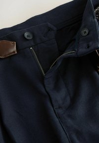 Pantalones azul marino con tejido texturizado, que presentan dos botones negros, cierre de cremallera y un detalle de cuero marrón cerca de la cintura.