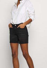 Pantalones cortos de mezclilla negra con un corte clásico, que cuentan con un diseño de cinco bolsillos y costuras visibles. Combinados con una camisa blanca de botones de ajuste holgado.