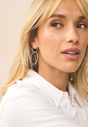 Blonde woman with light skin wears large silver hoop earrings and a white collared shirt, looking slightly to the right with lips parted.