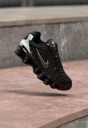 Chaussure de sport noire avec tige en mesh texturé, accents argentés, et une semelle épaisse et brillante noire comportant des éléments de rembourrage arrondis.