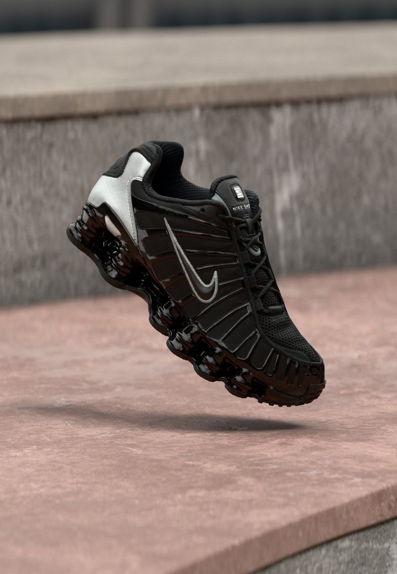 Zapato deportivo negro con parte superior de malla texturizada, detalles plateados y una suela negra gruesa y brillante con elementos acolchados redondeados.