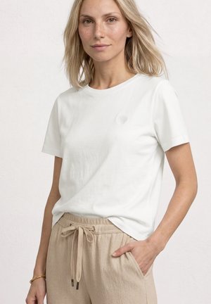 Blonde Frau trägt ein weißes kurzärmeliges Hemd mit Muschelemblem und beige Hose mit Kordelzug, eine Hand in der Tasche.