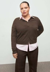 Brauner Cardigan mit Knöpfen, getragen über einem hellrosa Hemd, kombiniert mit braunen karierter Hose. Glatte Textur, figurbetontes Design.