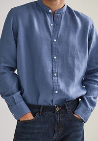 Ljusblå långärmad skjorta med ståkrage, knäppning framtill och manschetter, tillsammans med mörka denimjeans. Mjuk textur.