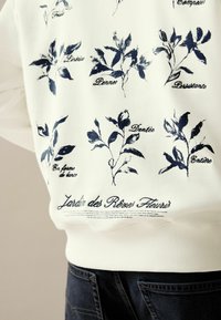 Bijela sweatshirt majica s tamnoplavim botaničkim uzorcima i tekstom, s rebrastim manžetama i donjim dijelom, koja prikazuje detaljan cvjetni dizajn.