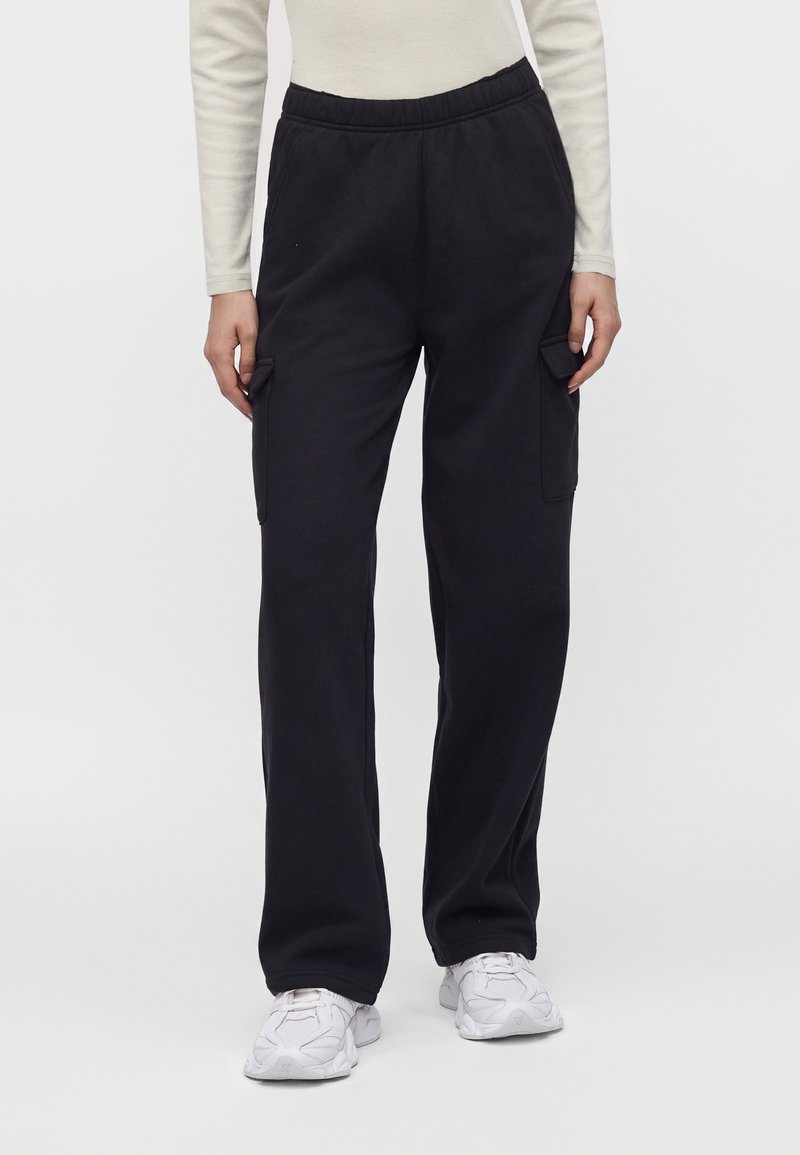 Personne portant un pantalon cargo noir avec une taille élastique et des poches à rabat, des baskets blanches et un haut clair à manches longues.