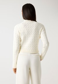 Maglione cream a trecce con un design aderente e orlo a coste. La parte posteriore presenta motivi a maglia testurizzati e maniche lunghe.