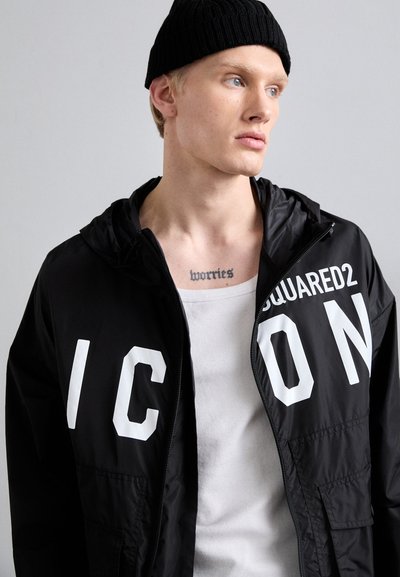Schwarze Kapuzenjacke mit oversized weißem Text "ICON" und "SQUARED2". Leichter Stoff mit glatter Textur. Getragen über einem weißen Tanktop.