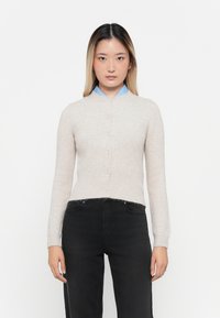 Cardigan de punto beige claro con cuello redondo, cierre de botones y puños acanalados. Llevado sobre una camisa de cuello azul y combinado con jeans negros.