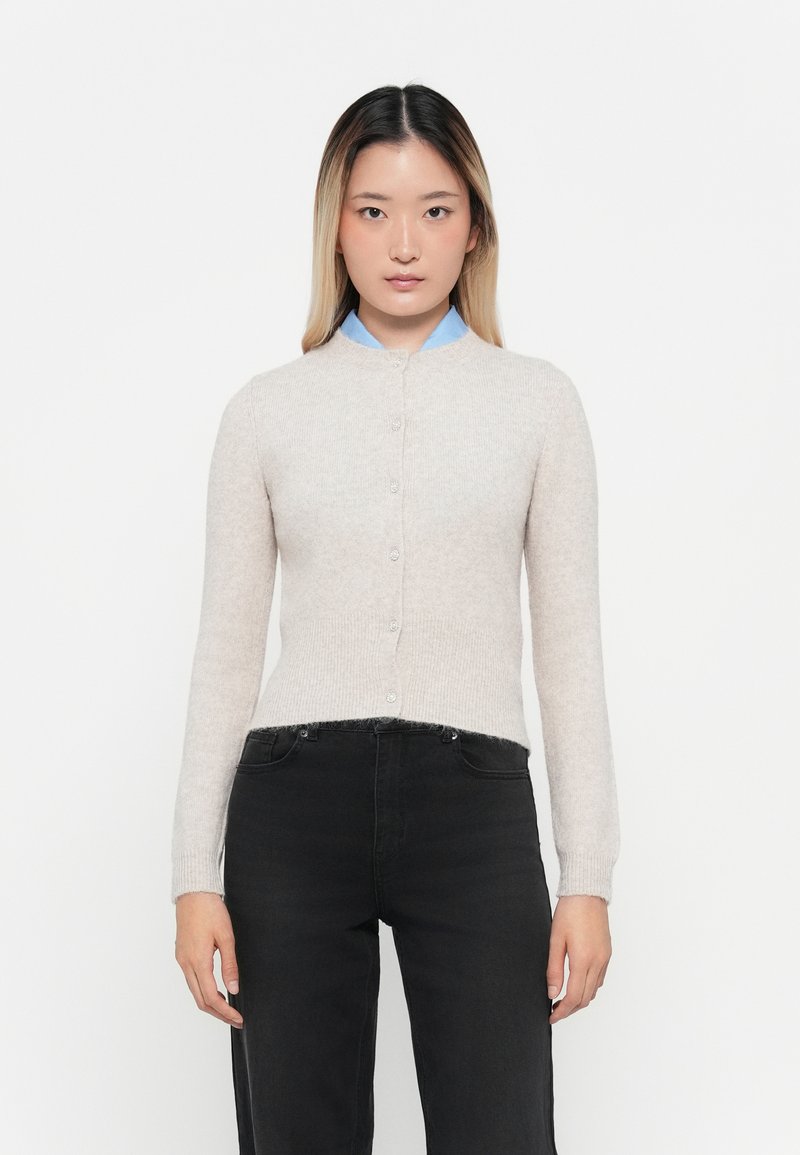 Cardigan de punto beige claro con cuello redondo, cierre de botones y puños acanalados. Llevado sobre una camisa de cuello azul y combinado con jeans negros.