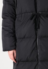 Svart dunjakke med glidelås, elastisk snøring i livet og horisontal quiltet tekstur. Har sidelommer og mansjetter på ermene.