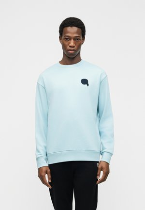 Młody mężczyzna w jasnoniebieskiej bluzie z niewielkim, ciemnym graficznym motywem dymka na piersi, stojący na tle jednolitego jasnego tła.