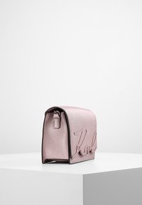 Vaaleanpunainen metallinhohtoinen käsilaukku, jossa on jämäkkä muoto, etupuolella kohokuvioitu "Karl"-kirjainkuvio ja hienovarainen tekstuuri.