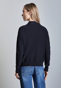 Marinblått stickat tröja med avslappnad passform, ribbade manschetter och fåll, kombinerad med blå denimjeans med dekorativa pärlor längs sidan.