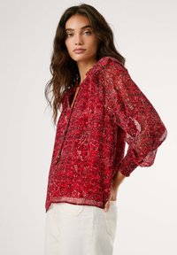 Blusa rossa a fantasia con un tessuto leggero e trasparente. Presenta un colletto rotondo, polsini aderenti e una vestibilità rilassata. Il design include motivi paisley intricati.