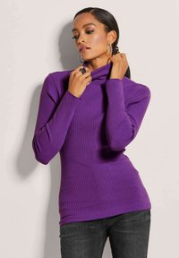 Lila gerippter Rollkragenpullover mit langen Ärmeln und taillierter Passform. Das Material wirkt weich mit einem sanften Glanz. Kombiniert mit dunkler Jeans.