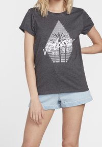 Grijze t-shirt met korte mouwen met een witte grafische afbeelding van palmbomen binnen een geometrische vorm. Gecombineerd met lichte denimshorts.