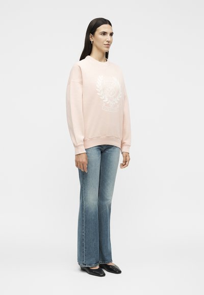 Femme aux longs cheveux foncés portant un sweat-shirt rose pâle avec un emblème blanc, un jean évasé bleu clair et des chaussures plates noires, debout de profil.