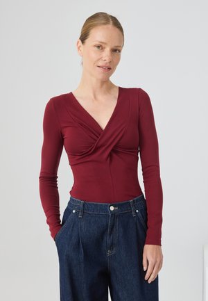 Body de manga larga burdeos con escote en V y diseño de frente retorcido, combinado con pantalones vaqueros oscuros de denim sueltos. Textura suave en todo el conjunto.