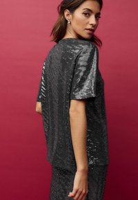 Top nero con paillettes, maniche corte, dal taglio morbido e texture lucida, su uno sfondo rosa.