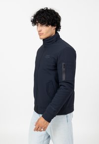 Marineblauer Bomberjacke mit hohem Kragen, Reißverschlusstaschen und gerippten Bündchen. Verfügt über ein dezentes Logo auf der Brust und einen strukturierten Stoff.