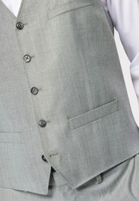 Grau-grüne Weste aus strukturiertem Stoff mit sechs Knöpfen, einer Brusttasche und kontrastierenden Nähdetails. Taillierte Passform.