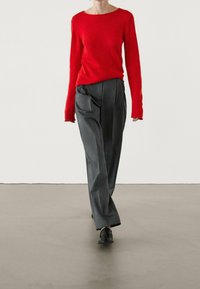 Un pull en tricot à manches longues rouge associé à un pantalon gris fuselé, avec des plis et une silhouette décontractée, porté avec des baskets noires.
