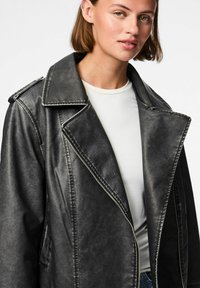 Veste en faux cuir noire avec un large col, fermeture éclair et coutures visibles. La texture alternative lui donne une apparence usée.