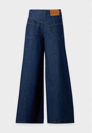 Mujer que lleva una camisa de manga corta a rayas beige y blanca, vaqueros de mezclilla azul oscuro de pierna ancha y zapatos marrones puntiagudos, de pie contra un fondo liso.