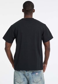 T-shirt en coton noir à manches courtes, col rond et coupe droite. Le tissu a une texture lisse. Porté avec un jean en denim bleu clair.