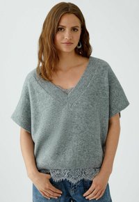 Frau in einem lockeren grauen V-Ausschnitt-Pullover über einem grauen Top mit Spitzenbesatz und blauen Jeans, die mit den Händen nahe den Taschen vor einem einfarbigen Hintergrund steht.