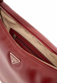 Sac à main en cuir synthétique rouge avec une fermeture éclair en ton doré. L'intérieur est doté d'une doublure crème avec l'imprimé "GUESS" et d'une petite poche zippée.