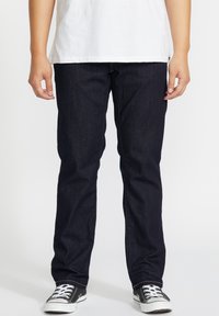 Vaqueros de mezclilla oscura con un corte recto, que presentan costuras visibles y bolsillos, combinados con zapatillas de lona negras y una camiseta blanca.