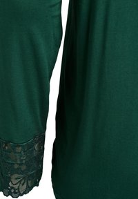 Temno zelena majica z čipkastimi poudarki na rokavih, mehko tkanino, sproščenim krojem in gladko teksturo s kančkom sijaja.