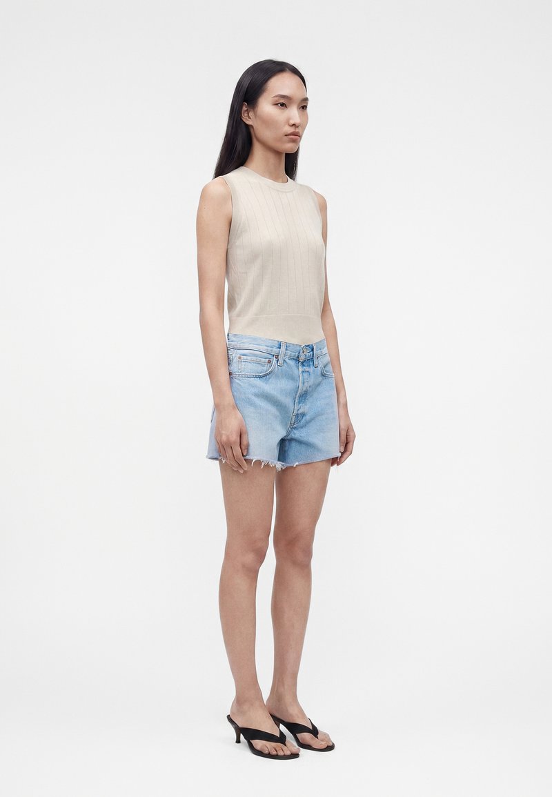 Jeune femme aux longs cheveux noirs, portant un haut beige sans manches, un short en jean bleu clair et des sandales à talons noires, debout devant un fond blanc.