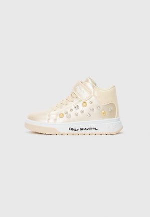 Cream high-top sneaker with gold and silver stud embellishments, velcro strap, and "Crazy Beautiful" text on the white sole.