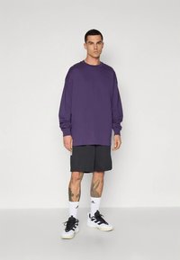 Purple long-sleeve shirt, loose fit, paired with black shorts. Model wears white sneakers with black accents and white socks. Simple background.