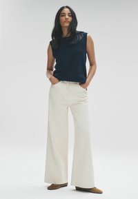 Top senza maniche blu navy abbinato a pantaloni a gamba larga color crema e scarpe in suede marroni, presentato su uno sfondo neutro.