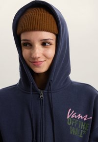 Marineblauwe hoodie met ritssluiting en een grote voorkantzak, met roze en groene logo's. Zachte textuur met een capuchon met trekkoord. Bruine geribbelde muts.