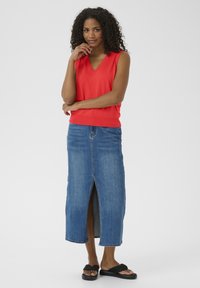 Un haut sans manches rouge avec un décolleté en V, associé à une jupe maxi en denim bleu présentant une fente sur le devant. Le modèle porte des sandales noires.