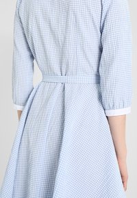 Kvinna som bär en ljusblå och vit rutig klänning med en åtsittande midja och trekvartslånga ärmar, stående med ryggen mot kameran.