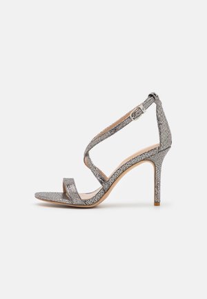 Sølvfarvet højhælet sandal med tynd ankelrem, åben tå og tekstureret mønster på overdel og hæl mod hvid baggrund.