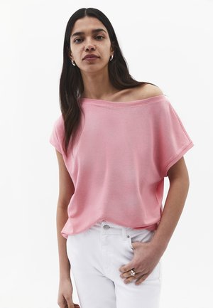Frau mit langen dunklen Haaren trägt ein lockeres rosa Off-Shoulder-Top und weiße Hosen, steht mit einer Hand in der Tasche vor einem schlichten Hintergrund.