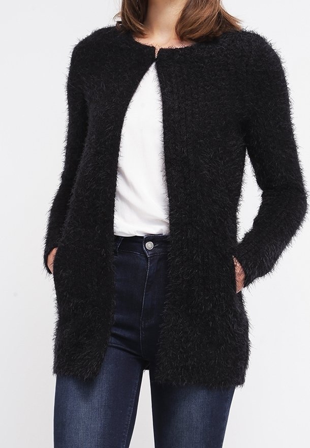 Femme portant un long cardigan noir en peluche par-dessus une chemise blanche et un jean bleu foncé, les mains partiellement dans les poches du cardigan.