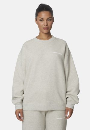 Donna che indossa una felpa oversize beige chiaro e pantaloni coordinati con piccolo testo bianco sul petto e sulla coscia, in piedi su sfondo bianco.