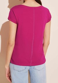 Fuchsie tričko s krátkým rukávem vyrobené z měkké bavlny, s detailem švu na zádech a ohrnutým rukávem. Kombinováno se světle modrými džíny.